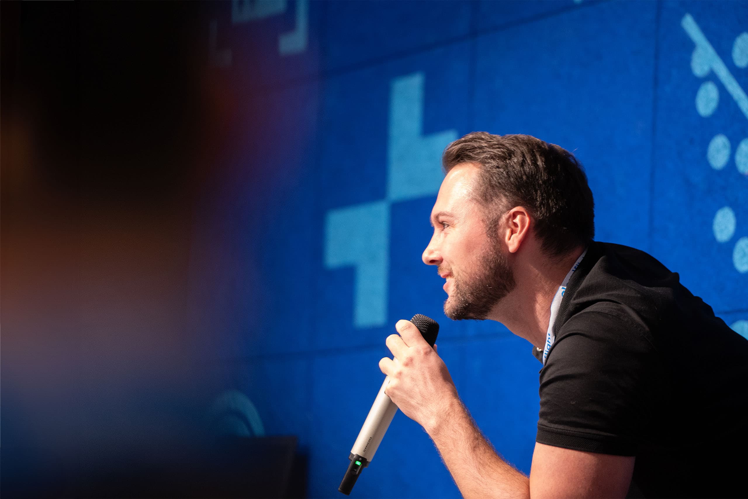 moderator with microphone on a blue stage, wearing a corporate lanyard with Cloudflight logo
